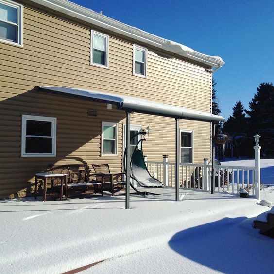 Aluminium patio awning with snow cover beside tan house, outdoor furniture under awning